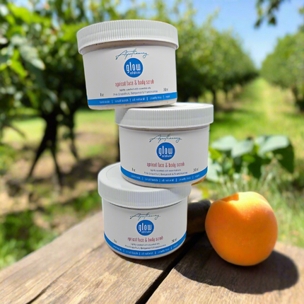 a stack of 3 jars of apricot face and body scrub and an apricot on a picnic table in an apricot orchard. 